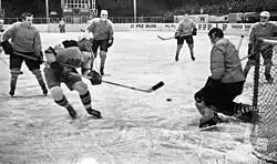 Black and white in-game action photo of a United States player shooting the puck at the net surrounded by Yugoslavia players