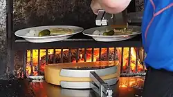 A half raclette cheese being melted on a wood fire while the other half is being scraped from its melted part in a plate