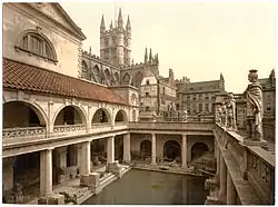 A late-nineteenth-century Photochrom of the Great Bath at the Roman Baths. Pillars tower over the water, and the spires of Bath Abbey&nbsp;– restored in the early sixteenth century&nbsp;– are visible in the background.
