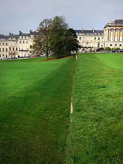 A ditch or trench in the grass with the right hand wall vertical and the left hand wall sloping. In the background are trees and yellow coloured stone buildings.