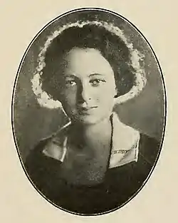 A young white woman with wavy dark hair, wearing a dark garment with a white collar; the lighting of the photograph lends a halo effect to her hair