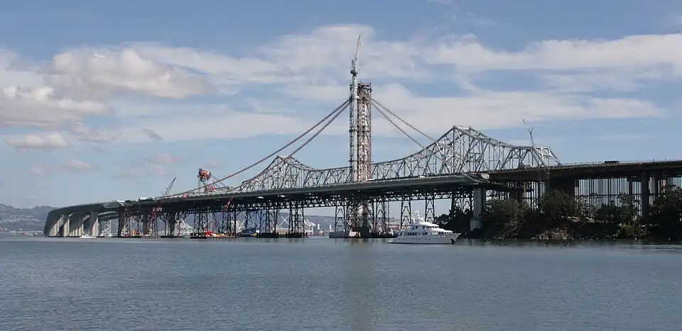 October 1, 2011: In the distance, the Left Coast Lifter is placing the last of the main span deck segments. Two additional short segments will join the main span to the curved skyway extension. The main suspension cables will follow the curves outlined by the recently installed catwalks. Ten holdback cables to the right of the tower (below the catwalks) preload the tower, bending it 17 inches (430&nbsp;mm) west against the forces to be imposed by the main cable when the bridge is complete, allowing the tower to be vertical when the holdbacks are removed. Subsequent to this image the traveler support cabling and cabling supports were installed, and all of the main cable strands have been placed and compacted, the suspender cables hung, attached, and tensioned.