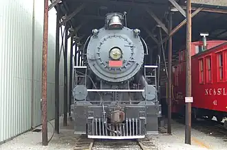 A black steam locomotive sitting in outdoor storage beneath a shelter, with a caboose on the right, and a shop building on the left