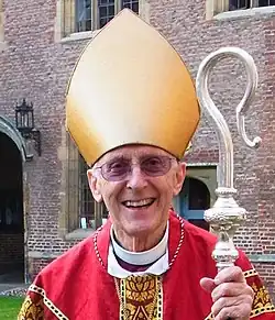 A smiling, bespectacled old man in a red chasuble and gold mitre, holding a crozier and standing on the lawn in front of a large red-brick building.