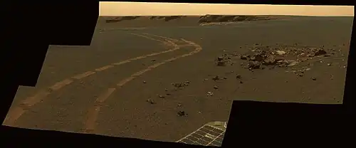 Figure 9. Sol 1162 (2007-05-01). The vicinity of the rim of Victoria Crater. This image shows a tiny (~1 m diameter) crater, Granada Crater, and this crater's ejecta in the right foreground, and a large 800m diameter crater, Victoria Crater, in the left background. The tracks of the rover Opportunity wind across the image around the two craters. The larger crater is much older (> 140 million years) than the smaller one (a few hundred thousand years) and in a more advanced state of erosion. An expanse of smooth sandy soils fill out the background. Image taken on Sol 1162 (2007-05-01).