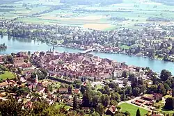 Aerial picture showing a small walled city dotted with red tile roofs and steeples; a wide river runs between it and another city. The farmlands on the far side are bathed in sunlight.