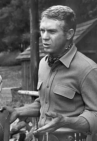 Black-and-white publicity photograph of a Caucasian man. The man has blonde hair and is wearing a shirt and a kerchief around the neck.