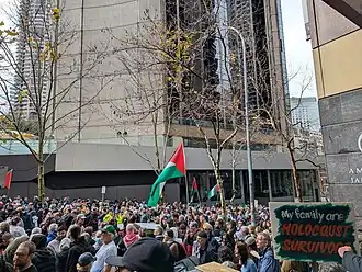 A photo of a crowd of protesters packed together. Multiple protesters have Palestinian flags and one has a sign that says "My family were Holocaust survivors". They are in front a tall, city building with another visible in the background. There are four mostly bare trees with a few autumn leaves.