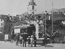 Black and white photo of a horse pulling an enclosed four-wheel tramcar and a smaller two-wheel trailer for transporting post.