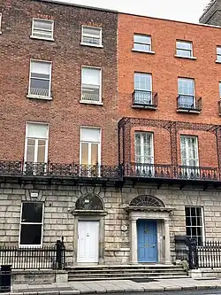 This image is of the Royal Institute of the Architects of Ireland on Merrion Square. It is a four-story building with two doors next to each other one with a blue door and one with a white door. There is a silver sign that says RIAI beside the door.