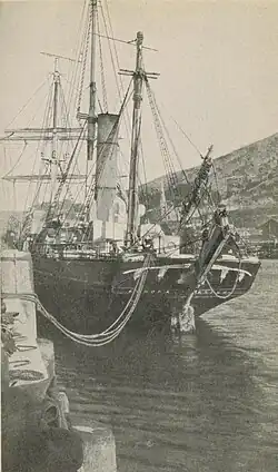 A ship with three masts and a tall central funnel, tied to the dockside with loose ropes so that the stern is swinging outwards