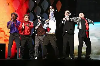 Six men wearing multicolored clothes are seen performing onstage.