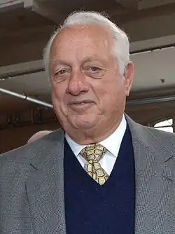 A man in a gray suit jacket, navy sweater, white shirt, and gold tie