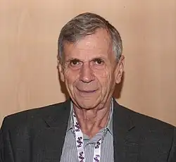 An older man in a dress suit standing in front of a wood background.