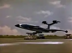 A gleaming, blue, futuristic airliner rests on top of three orange support vehicles (one underneath the fuselage and two underneath the wings) as it speeds out of control along a runway against a grassy backdrop, the sky bright with clouds.