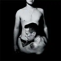 A black-and-white photo of two shirtless men. Both are wearing necklaces with a cross on it; one is holding the other by the torso.