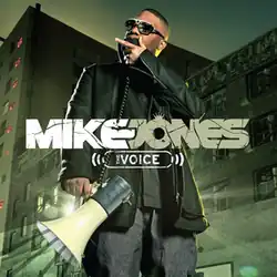 The cover shows a man wearing a black jacket, grey t-shirt, blue jeans and sunglasses in front of an apartment building. He's holding and speaking into a megaphone with light shining behind it.
