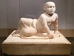 Beige marble statue of a stout young child aged about two years old lying on his left side. The child's head is shaven, his eyes gaze over the viewer's shoulder and his lower body is covered in a draping cloth that hangs limply between his flexed feet. The child supports his torso with his left hand in which he holds an unidentifiable object, he also holds a small bird in his right hand. The sculpture rests on a heavy socle inscribed with barely visible letters spanning the upper part of the socle vertically.
