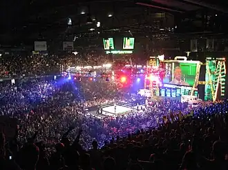 A sold out crowd watching the event, with a professional wrestling ring in the center of the picture. A giant screen in the shape of a briefcase is at the top right.