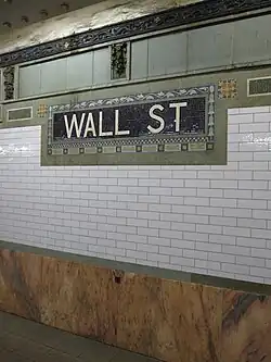 View of the wall along the platform. There is a a pink marble wainscoting on the lowest part of the wall, with white glass tiles above. A white-on-blue tile plaque with the words "Wall Street" and floral motifs is also placed on the wall.