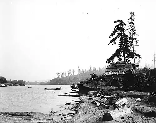 A black and white photo of an old plank house belonging to Salmon Bay Charlie along the edge of the bay, with a Shilshole canoe moored outside