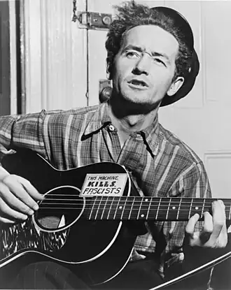 Black-and-white photo of Guthrie looking up at an angle away from the camera and playing a guitar that says "This machine kills fascists"