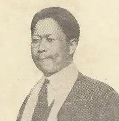 A black-and-white, slightly yellowed, newspaper scan of a man in a suit and tie, wearing glasses. He has short black hair and has a slightly unpleased expression.