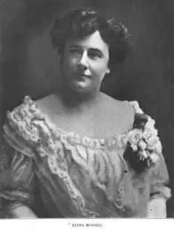 A white woman with dark hair in a bouffant updo, wearing a white frilly dress with a wide scooped neckline and a corsage