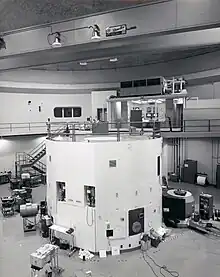 The black and white photograph is of a large room that contains a lot of electronic equipment. The lower half of the image contains a cylindrical white container that is a nuclear reactor. There is a walkway at the top of the reactor, which leads back to a control room where two men are sitting.
